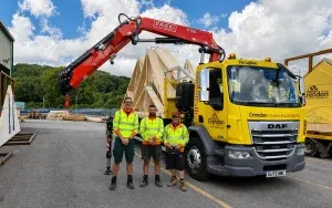 Three workers in front of a yellow lorry holding roof trusses with a crane attached
